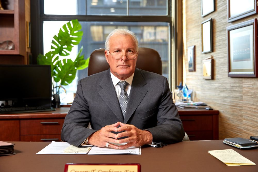 Professional businessman in suit seated at office desk, looking confidently at camera with hands folded.