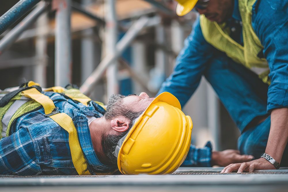 Construction worker down on site, receiving aid from colleague in safety gear.