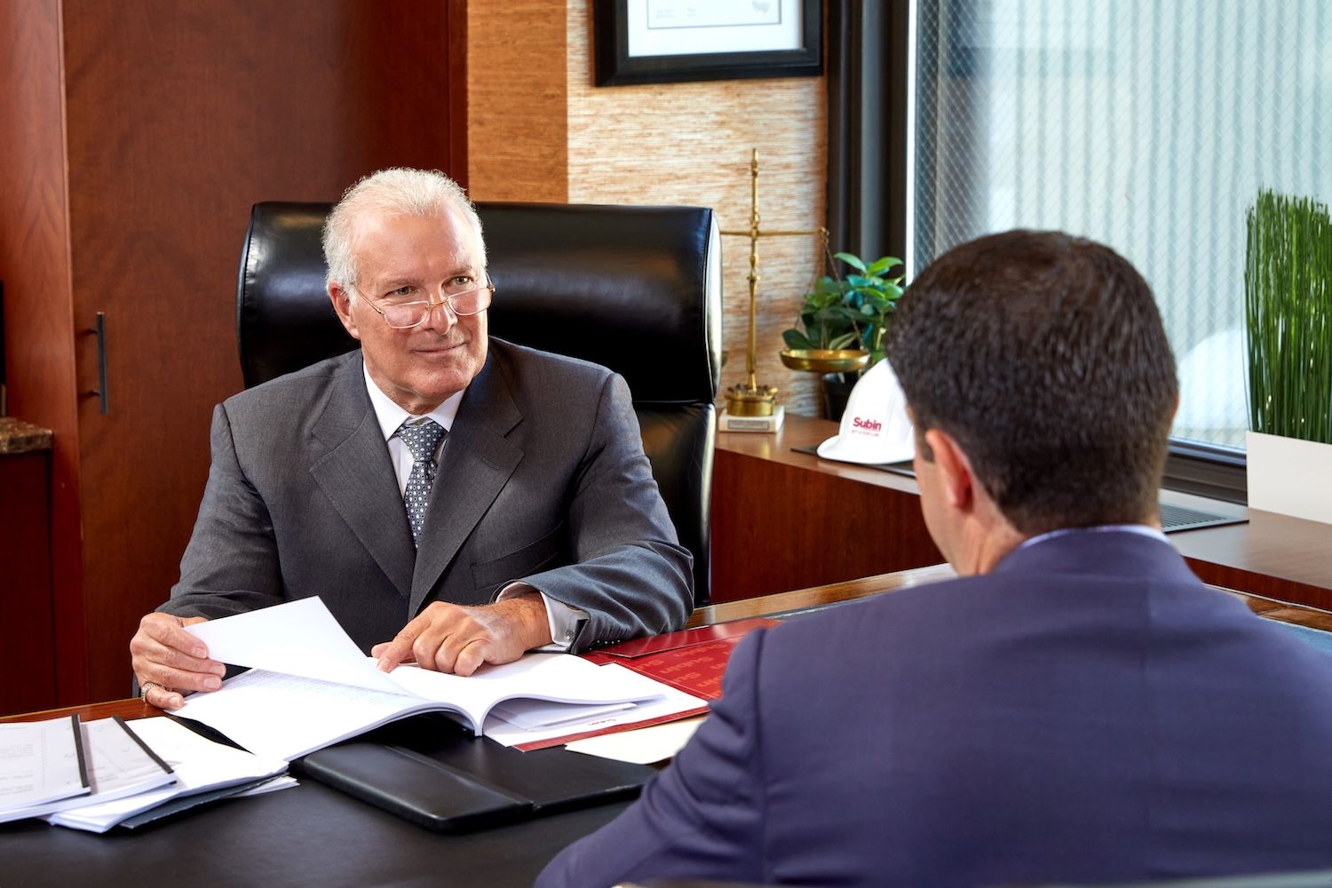 Two businessmen in suits having a discussion at an office desk with documents and hard hat nearby.