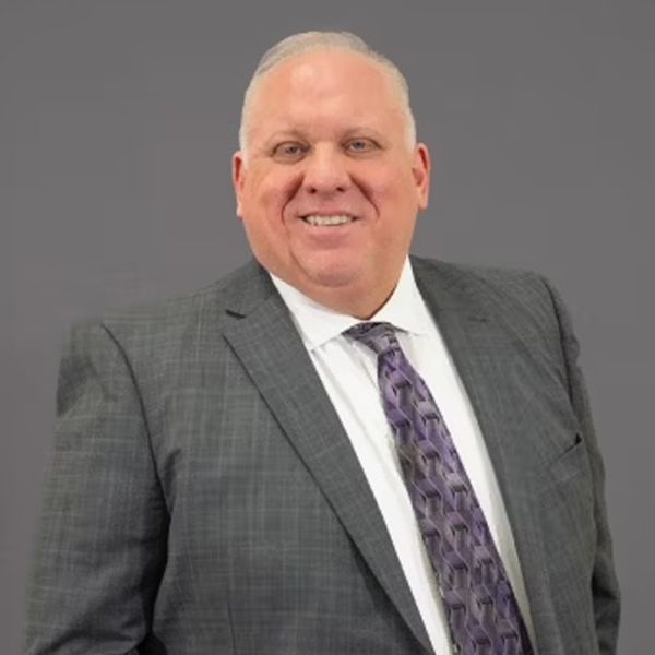 Smiling man in a suit with a purple tie stands against a gray background.