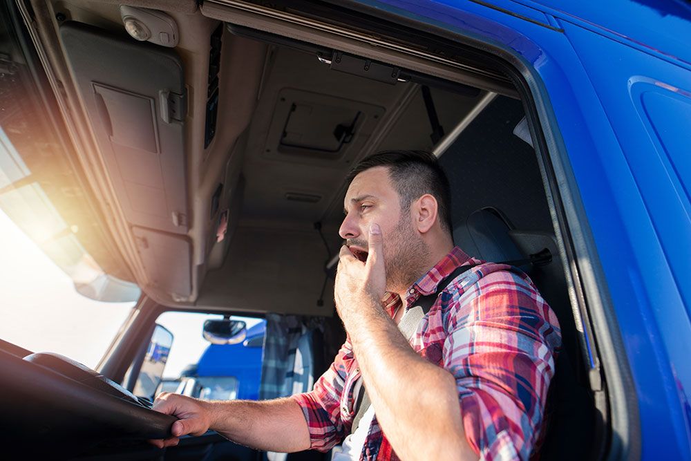 Truck driver in a blue cab yawning, illustrating fatigue or tiredness during a long-distance trip.