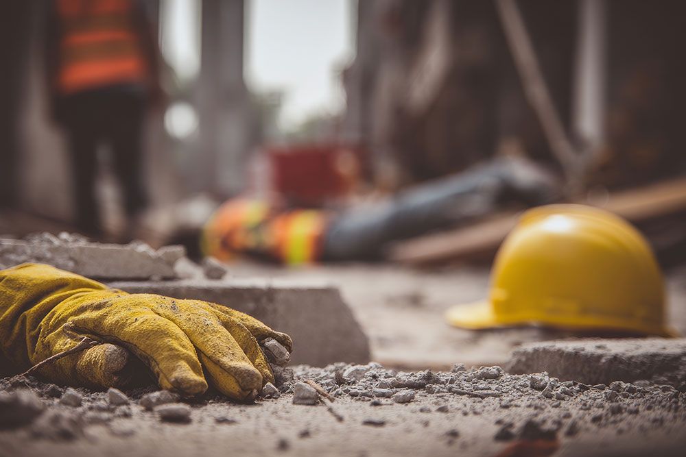 Construction site with yellow glove and helmet in focus, worker blurred in background. Safety equipment emphasized.