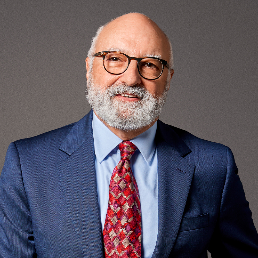 Elderly man with glasses, white beard, and a blue suit smiling against a gray background.