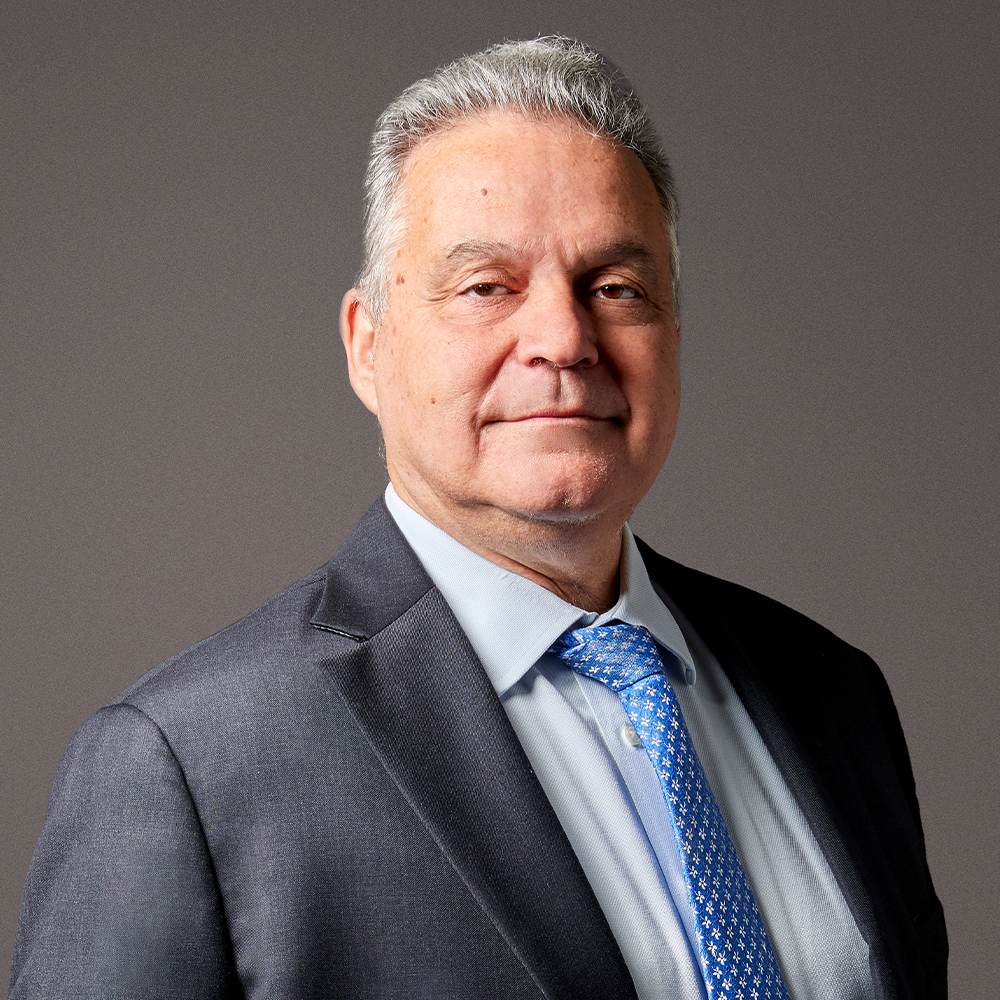 Senior man in a suit and blue tie against a gray background, facing slightly to the side.